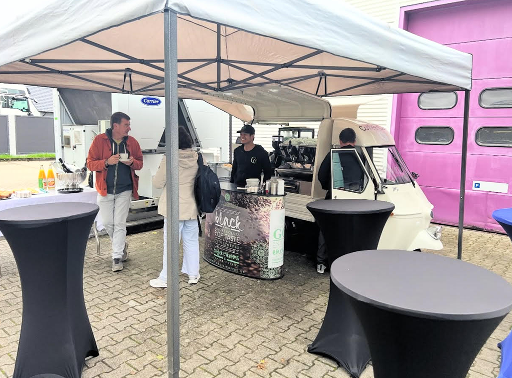 Mobile Kaffeebar unter Pavillon bei einem Firmenevent, Gäste trinken Heißgetränke an schwarzen Stehtischen.